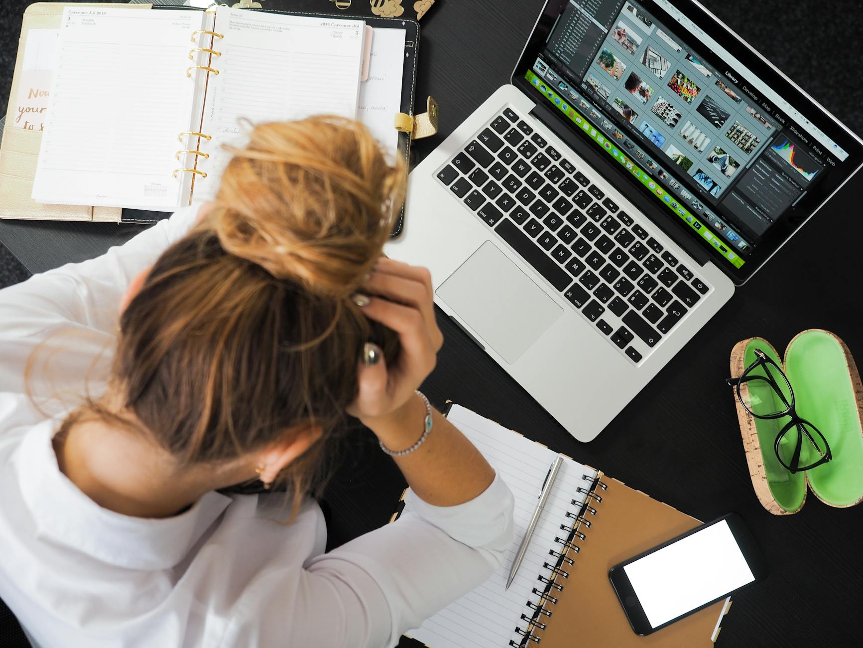 A woman is sitting at a table with her head in her hands. A daily planner is open next to her along with a notebook, her phone and a laptop with information on the screen.